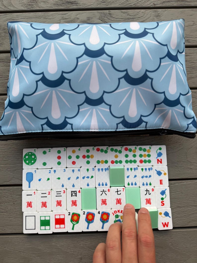 May include: A Mahjong set with white tiles featuring Chinese characters and colorful symbols. The set includes green tiles and is displayed on a wooden surface. A light blue zippered pouch with a scalloped pattern is in the background.