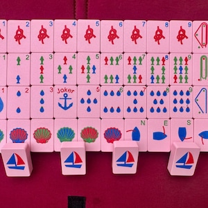 May include: A set of pink Mahjong tiles with various symbols and numbers, including anchors, boats, and water droplets. The tiles are arranged on a red surface, with four white boat-shaped tile holders.
