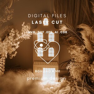 Op de afbeelding: Een lasergesneden digitale bestand van een huisvormig ontwerp met een hart- en bloemmotief. Het ontwerp bevat de tekst "DIGITAL FILES LASER CUT" en bestandstype afkortingen. Het huis heeft meerdere ramen en de tekst "BOHOHO ME" en "premium design".