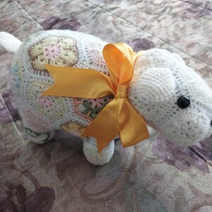 May include: A white crocheted stuffed animal shaped like a sheep with a yellow bow. The sheep has a crocheted body with a white and multi-colored pattern and a crocheted head with black eyes.