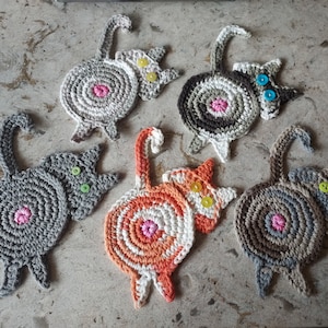May include: Set of six crocheted cat-shaped coasters in various colors, including gray, orange, and brown. Each coaster features a different colored button for the cat's eye.