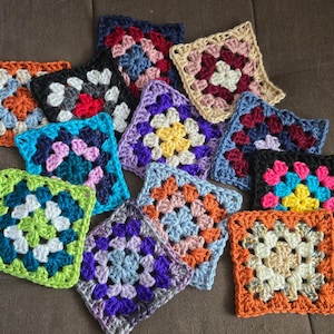May include: A collection of twelve square crochet granny squares in various color combinations including orange, blue, purple, yellow, pink, black, and white. Each square features a central flower-like pattern with concentric rows of yarn stitches.