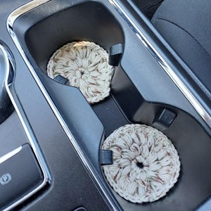 May include: Two crocheted cup coasters in a car cup holder. The coasters are beige with brown and green accents.