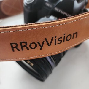 May include: A brown leather camera strap with the text "RRoyVision" embossed in black.