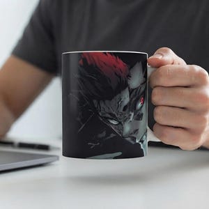 以下が含まれることがあります： 白いセラミック製のマグカップ。トゲのある赤い髪、鋭い顔立ち、そして深く暗い背景に浮かび上がる一つだけ光る赤い目をしたアニメキャラクターの、コントラストの強いダークなグラフィックイラストが描かれています。