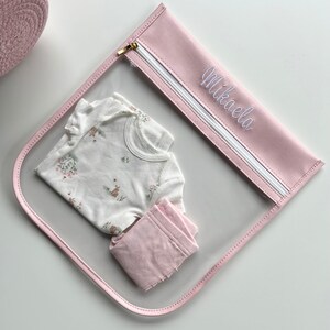 May include: A clear plastic pouch with pink trim and a gold zipper, featuring the name Mikaela embroidered in white script. Inside the pouch is a white short-sleeved onesie with a small floral and animal print, paired with folded light pink pants.