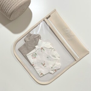May include: A clear plastic storage pouch with a beige trim and a zipper closure featuring the word Baby embroidered in cursive script. Inside the pouch are two folded garments: a solid taupe short-sleeve bodysuit and a white short-sleeve bodysuit with a small floral and animal print pattern.