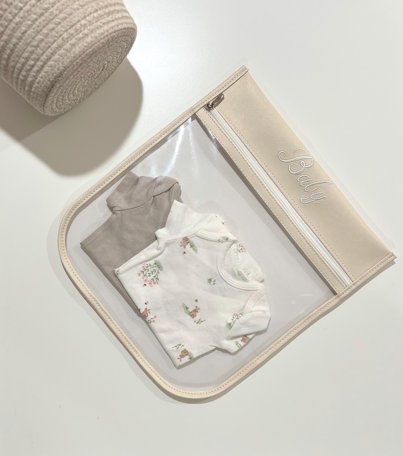 May include: A clear plastic storage pouch with a beige trim and a zipper closure featuring the word Baby embroidered in cursive script. Inside the pouch are two folded garments: a solid taupe short-sleeve bodysuit and a white short-sleeve bodysuit with a small floral and animal print pattern.