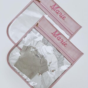 May include: Two clear plastic storage pouches with dusty pink trim and gold zippers. Each pouch features the name Marie embroidered in pink thread. Inside the pouches are folded baby clothes, including a tan striped bodysuit and white shirts.