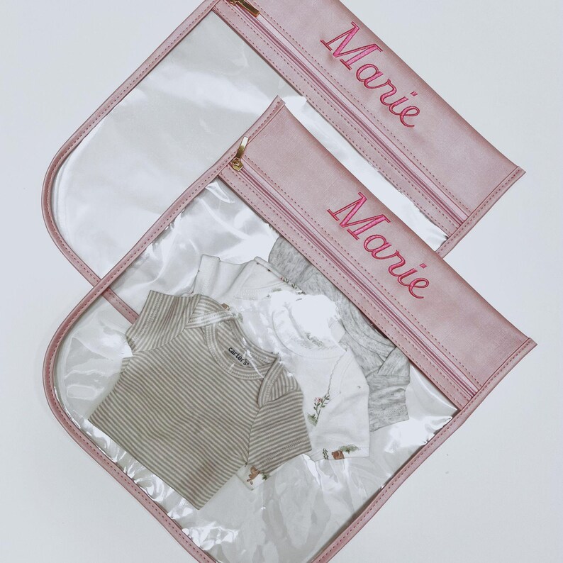 May include: Two clear plastic storage pouches with dusty pink trim and gold zippers. Each pouch features the name Marie embroidered in pink thread. Inside the pouches are folded baby clothes, including a tan striped bodysuit and white shirts.