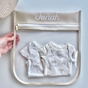 May include: A clear plastic storage pouch with a cream-colored trim and zipper, featuring the name Jonah embroidered in white script. Inside the pouch are two short-sleeved baby bodysuits, one light gray and one cream with a small animal print.