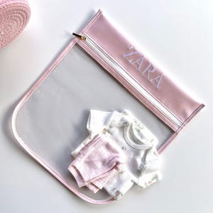 May include: A pink baby travel bag with a clear front, a gold zipper, and the name "ZARA" embroidered in white. Inside the bag are a white floral onesie and pink baby pants.
