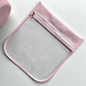May include: A square, clear plastic pouch with a light pink fabric border and a matching pink top panel. The top panel features the name Mikaela embroidered in white script. A white zipper with a gold-toned metal pull runs across the top.