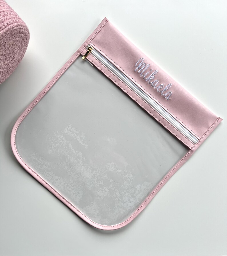 May include: A square, clear plastic pouch with a light pink fabric border and a matching pink top panel. The top panel features the name Mikaela embroidered in white script. A white zipper with a gold-toned metal pull runs across the top.