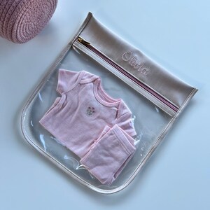 May include: A clear plastic storage pouch with a beige fabric top featuring the embroidered name Olivia and a gold zipper. Inside the pouch is a light pink short-sleeved baby bodysuit with a small floral embroidery on the chest and matching pants.