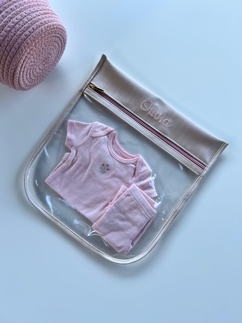 May include: A clear plastic storage pouch with a beige fabric top featuring the embroidered name Olivia and a gold zipper. Inside the pouch is a light pink short-sleeved baby bodysuit with a small floral embroidery on the chest and matching pants.