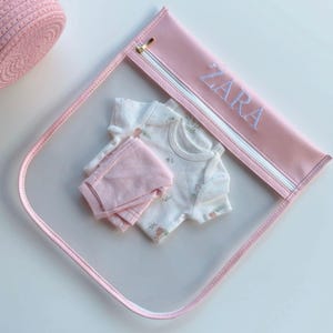 May include: A pink, clear, zippered pouch with the name "ZARA" embroidered in white. Inside the pouch are a white onesie with a floral pattern and a pair of pink shorts. A pink woven basket is in the upper left corner.