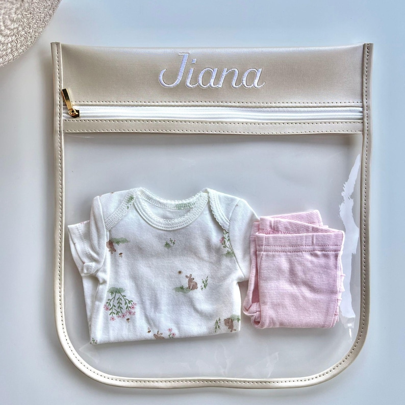 May include: A clear plastic storage pouch with a beige trim and a gold zipper, featuring the name Jiana embroidered in white thread. Inside the pouch is a white short-sleeved bodysuit with a small floral and rabbit print and light pink leggings.