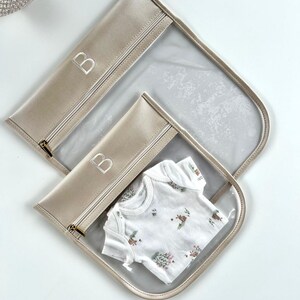May include: Two clear storage pouches with beige trim and gold-tone zippers, each featuring the letter B embroidered in white. The smaller pouch contains a white short-sleeved garment with a small floral and animal print pattern in muted tones.