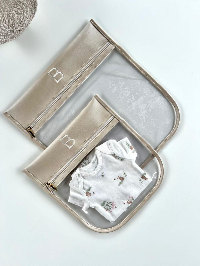 May include: Two clear storage pouches with beige trim and gold-tone zippers, each featuring the letter B embroidered in white. The smaller pouch contains a white short-sleeved garment with a small floral and animal print pattern in muted tones.