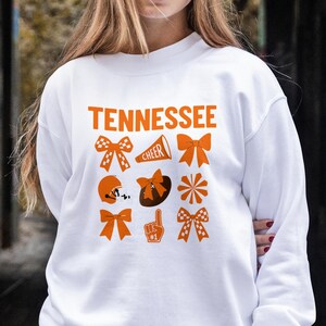 Może przedstawiać: Biała bluza z napisem "Tennessee" w pomarańczowych literach. Poniżej pomarańczowe grafiki kokard, kasku do futbolu amerykańskiego, pomponu i piankowego palca. Noszona przez osobę w dżinsach.