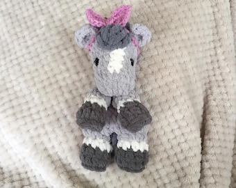 May include: A handmade crochet horse doll in shades of gray, white, and dark gray. The horse has a white patch on its face and a pink bow. The doll is made of soft yarn and is a cute, cuddly toy.