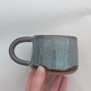 May include: A ceramic mug with a rounded handle, featuring a mottled blue and grey glaze on the upper portion and a solid brown base. The mug has a cylindrical shape with a smooth rim and a textured, speckled finish across the exterior surface.