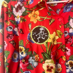 May include: Red button-down shirt with a vibrant floral pattern featuring pansies and other flowers. A circular patch with a white figure is sewn on the front. The shirt has a collar and long sleeves. The brand name "Linea by Louis Dell'Olio" is visible.
