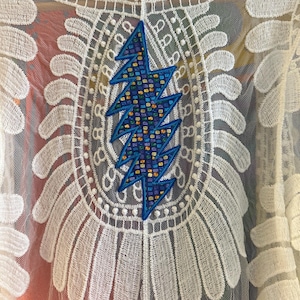 May include: White lace top with a large, blue lightning bolt design on the back. The lightning bolt is filled with a colorful mosaic pattern. The top features intricate lace detailing around the neckline and sleeves, creating a delicate and bohemian aesthetic.