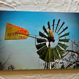 May include: A vintage Aermotor windmill with a yellow and red sign that reads "Aermotor USA" against a blue sky. The windmill has a large, circular frame with multiple blades.