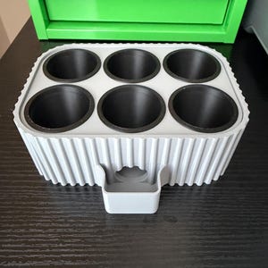 May include: A light gray, 3D-printed organizer with six circular compartments and a small handle. The organizer is designed to hold small items. The background includes a green toolbox and a black surface.