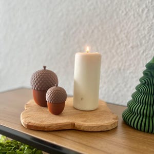 Könnte beinhalten: Eine brennende weiße Säulenkerze und zwei eichelförmige Dekorationen stehen auf einem Holztablett. Die Eicheln sind braun und orange, und das Tablett steht auf einem Holzregal. Rechts ist ein grüner, stilisierter Weihnachtsbaum.