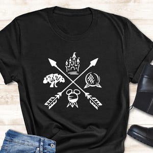 May include: Black t-shirt with a white graphic featuring crossed arrows, a castle, a tree, a globe, and a Mickey Mouse head. The shirt is paired with blue jeans, sunglasses, and black ankle boots. A small potted plant is also present.