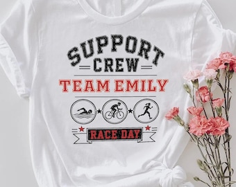 May include: A white short-sleeved t-shirt featuring black and red graphic text that reads SUPPORT CREW TEAM EMILY RACE DAY. The design includes three circular icons depicting a swimmer, a cyclist, and a runner, accented with small red stars.