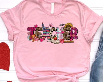 May include: Pink t-shirt with the word "TEACHER" spelled out in colorful patterned letters. The letters are decorated with various designs, including hearts, flowers, and a ladybug. The shirt is displayed on a white background with a pair of blue jeans and red hearts.