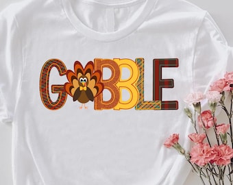 May include: White t-shirt with the word "GOBBLE" in colorful, patterned letters. The letter "O" is replaced with a turkey illustration. The shirt is styled with a knot at the bottom and a bouquet of pink flowers.