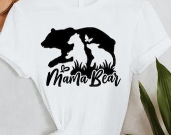 May include: White t-shirt with a black graphic of a bear, two cubs, and a butterfly, with the words "Mama Bear" in a script font. A tan hat hangs on the wall behind the shirt. Green plant leaves are visible on the right.