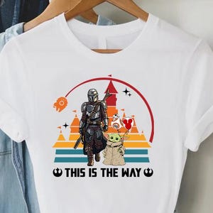 May include: White t-shirt featuring a graphic of The Mandalorian and Grogu, set against a sunset backdrop with a castle silhouette. The text "THIS IS THE WAY" is printed below the image. The shirt is hanging on a wooden hanger.
