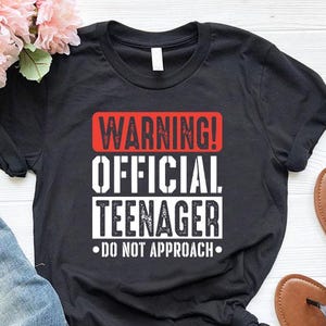 May include: Black t-shirt with the text "WARNING! OFFICIAL TEENAGER - DO NOT APPROACH" in white and red. The shirt is paired with blue jeans and brown sandals. Pink flowers are in the background.