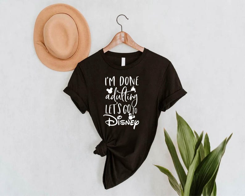 May include: Black t-shirt with the text "I'M DONE adulting LET'S GO TO Disney" in white script. The shirt is hanging on a wooden hanger. A tan hat is visible in the background.