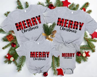 May include: Four gray t-shirts and a onesie with the text "MERRY Christmas" in red and black plaid. Festive holiday apparel with Christmas decorations, including red ornaments and greenery.