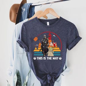 May include: Navy blue t-shirt featuring a graphic of The Mandalorian and Grogu, with a red and white heart-shaped balloon. The design includes a sunset and the text "THIS IS THE WAY".