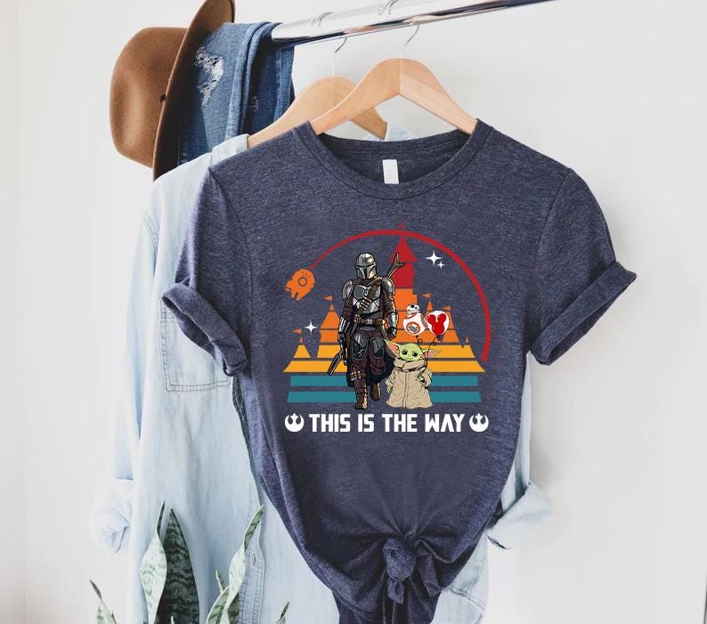 May include: Navy blue t-shirt featuring a graphic of The Mandalorian and Grogu, with a red and white heart-shaped balloon. The design includes a sunset and the text "THIS IS THE WAY".