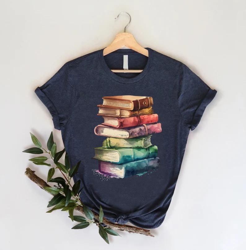 May include: Navy blue t-shirt featuring a watercolor illustration of a stack of books. The books are in various colors, including brown, red, green, and blue. The shirt is hanging on a wooden hanger against a white background.