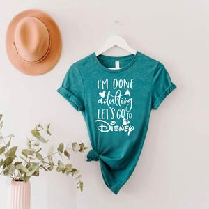 May include: Teal t-shirt with the text "I'm done adulting let's go to Disney" in white script. The shirt is hanging on a white hanger. A tan hat is on the wall above the shirt. A vase with greenery is in the lower left corner.
