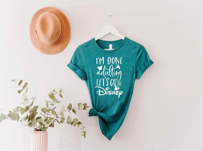 May include: Teal t-shirt with the text "I'm done adulting let's go to Disney" in white script. The shirt is hanging on a white hanger. A tan hat is on the wall above the shirt. A vase with greenery is in the lower left corner.