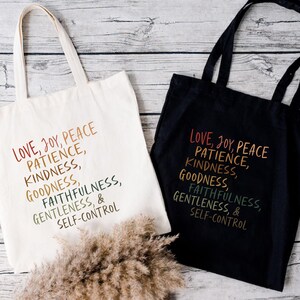 Pode incluir: Duas sacolas, uma creme e outra preta, com as palavras "LOVE, JOY, PEACE, PATIENCE, KINDNESS, GOODNESS, FAITHFULNESS, GENTLENESS, & SELF-CONTROL" impressas em um gradiente de arco-&iacute;ris. As sacolas t&ecirc;m al&ccedil;as longas.