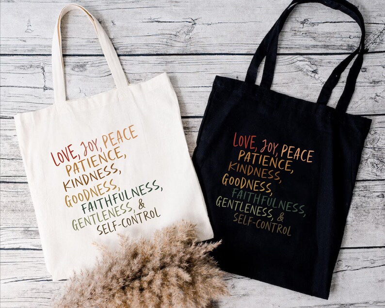 Pode incluir: Duas sacolas, uma creme e outra preta, com as palavras "LOVE, JOY, PEACE, PATIENCE, KINDNESS, GOODNESS, FAITHFULNESS, GENTLENESS, & SELF-CONTROL" impressas em um gradiente de arco-&iacute;ris. As sacolas t&ecirc;m al&ccedil;as longas.