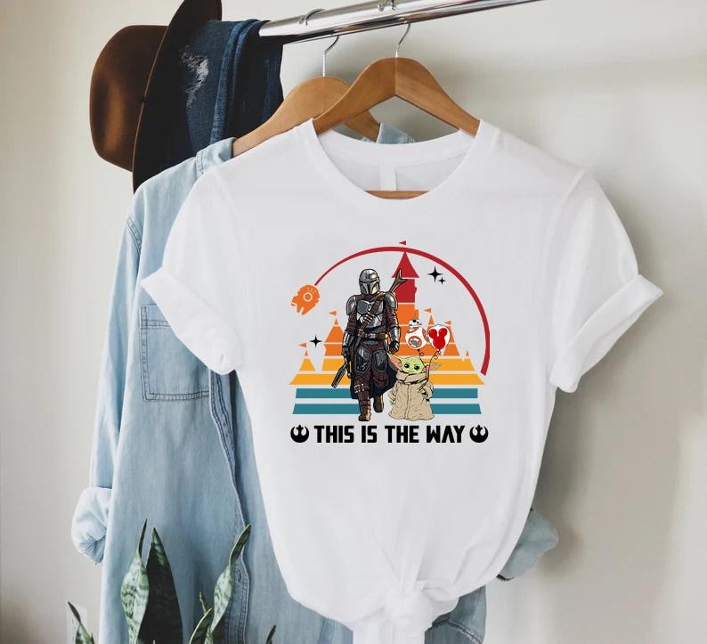 May include: White t-shirt featuring a graphic of The Mandalorian and Grogu, set against a sunset backdrop with a castle silhouette. The text "THIS IS THE WAY" is printed below the image. The shirt is hanging on a wooden hanger.