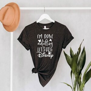 May include: Dark gray t-shirt with the text "I'M DONE adulting LET'S GO to Disney" in white script. The shirt is hanging on a white hanger. A brown hat hangs on a white pole to the left.